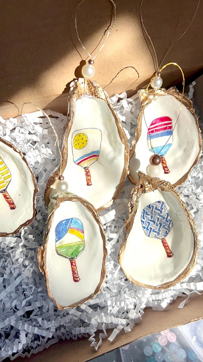 May include: Five oyster shell ornaments with gold trim, each featuring a different hand-painted design of a paddleball racket. The rackets are painted in various colors and patterns, including yellow, blue, red, and green.