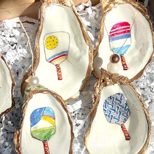 May include: Five gold-rimmed oyster shell ornaments with different colored paddleball rackets painted inside. Each ornament has a white pearl hanging from it.