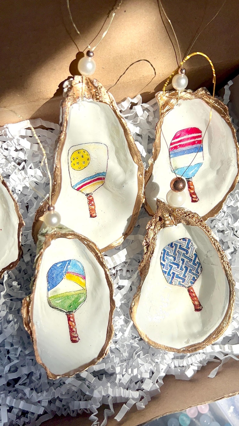 May include: Five gold-rimmed oyster shell ornaments with different colored paddleball rackets painted inside. Each ornament has a white pearl hanging from it.