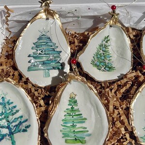 May include: Set of four oyster shell Christmas ornaments with gold trim and painted watercolor Christmas trees. Each ornament features a different design of a Christmas tree with a star on top.