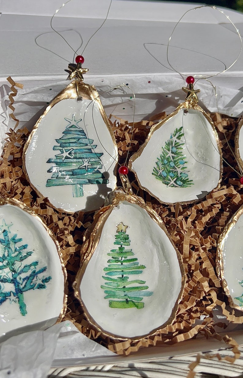 May include: Set of four oyster shell Christmas ornaments with gold trim and painted watercolor Christmas trees. Each ornament features a different design of a Christmas tree with a star on top.