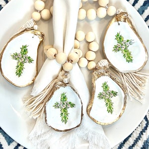 Könnte beinhalten: Ein Set aus vier Serviettenringen aus Austernmuscheln mit einem grünen Kreuzdesign. Die Ringe sind mit Goldfarbe verziert und haben jeweils einen weißen Quastenanhänger. Die Ringe werden auf einem weißen Teller mit einer Holzperlenkette präsentiert.