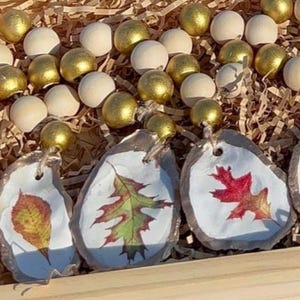 K&ouml;nnte beinhalten: Vier Herbst-Ornamente mit bemalten Bl&auml;ttern auf wei&szlig;em Hintergrund. Die Ornamente sind an einer Holzperlenkette mit goldenen und wei&szlig;en Perlen befestigt.