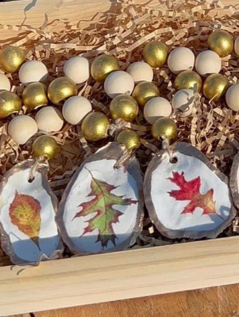 K&ouml;nnte beinhalten: Vier Herbst-Ornamente mit bemalten Bl&auml;ttern auf wei&szlig;em Hintergrund. Die Ornamente sind an einer Holzperlenkette mit goldenen und wei&szlig;en Perlen befestigt.