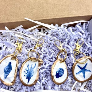 May include: Four handmade keychains with gold-colored hardware, each featuring a white oyster shell design with blue painted details. The shells have a gold-colored trim. The keychains are displayed in a cardboard box with white shredded paper.