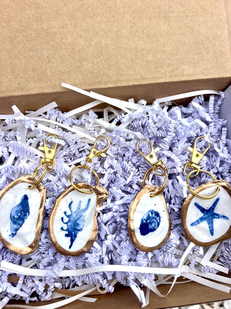 May include: Four handmade keychains with gold-colored hardware, each featuring a white oyster shell design with blue painted details. The shells have a gold-colored trim. The keychains are displayed in a cardboard box with white shredded paper.