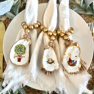 Op de afbeelding: Drie witte servetten met gouden kralen-servetringen en oester-placecardhouders. De oesterschelpen zijn versierd met gouden verf en hebben elk een ander ontwerp. De ontwerpen omvatten een boom met vogels, twee vogels en een hen met kuikens.