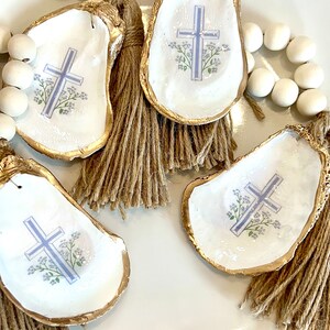 May include: Four oyster shells with gold trim and a blue cross with white flowers on a white plate. Each shell has a brown tassel.
