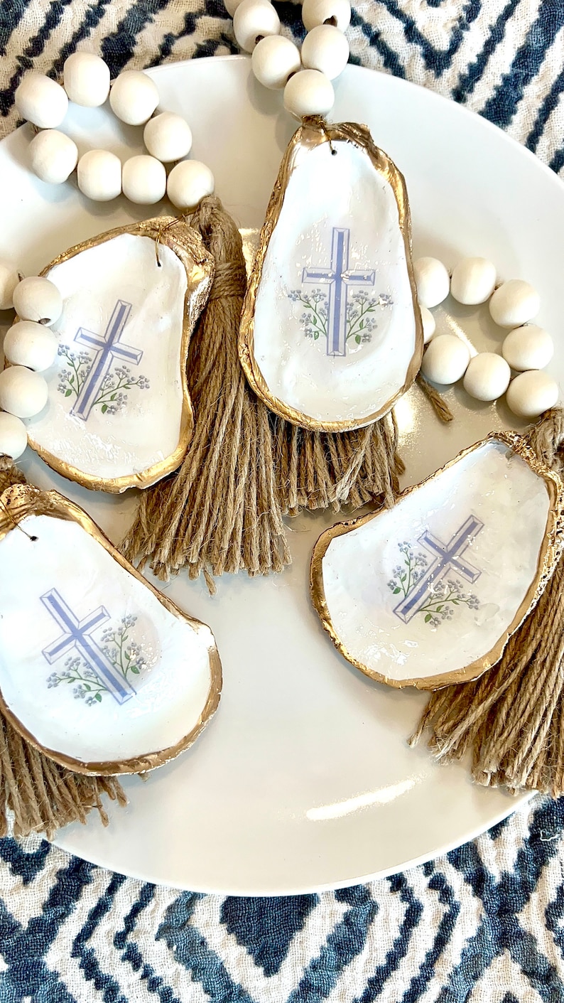 May include: Four oyster shells with gold trim and a blue cross with white flowers on a white plate. Each shell has a brown tassel.