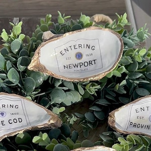 Pode incluir: Três pratos de lembrança de conchas de ostra brancas com acabamento dourado. Cada prato tem um rótulo preto e branco com o texto "Entering" e o nome de uma cidade: "Cape Cod", "Newport" e "Provincetown".