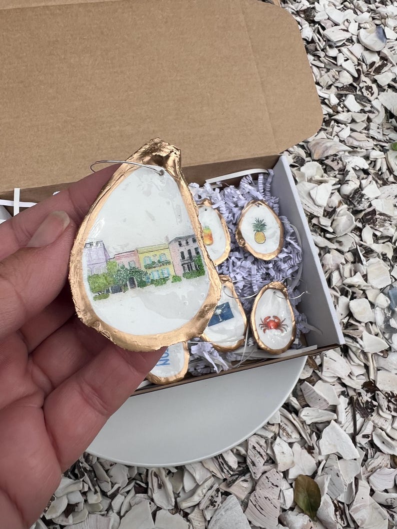 May include: A set of decorative oyster shell ornaments with gold trim. One shell features a watercolor painting of a street scene, while others display a pineapple, crab, and other designs. The shells are presented in a cardboard box.
