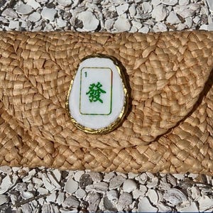 以下が含まれることがあります： フラップ開閉の織り込まれたストロークラッチ。 金色のフレームに囲まれた、緑色の漢字と数字の1が入った白いタイルの装飾が特徴です。 背景は白い貝殻の表面です。