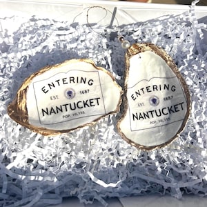 May include: Two white oyster shell ornaments with gold trim. Each ornament features a white sign with black text that reads "Entering Est. 1687 Nantucket Pop. 10,155".