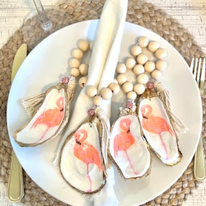 Op de afbeelding: Vier servetringen van beschilderde oesterschelpen met roze flamingo's, servetringen van houten kralen en witte kwastjes.