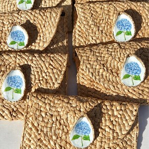 May include: A collection of rectangular woven straw clutch bags, each featuring a single decorative oyster shell attached to the front flap. Each shell is hand-painted with a blue hydrangea flower with green leaves on a white background.