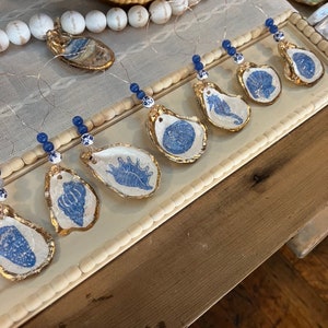 May include: A set of eight blue and white oyster shell ornaments with gold accents. Each ornament features a different sea creature design, including a seahorse, a seashell, and a starfish. The ornaments are strung on white twine with blue beads.