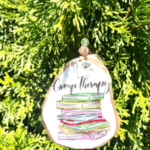 May include: A white ceramic ornament shaped like a book stack with the text "Group Therapy" on it. The ornament is decorated with colorful illustrations of books and has a gold string for hanging.