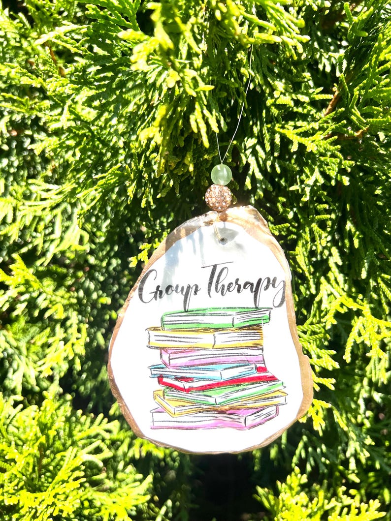 May include: A white ceramic ornament shaped like a book stack with the text "Group Therapy" on it. The ornament is decorated with colorful illustrations of books and has a gold string for hanging.