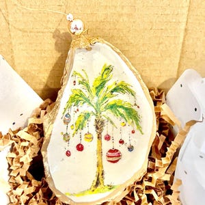May include: A decorative oyster shell ornament featuring a hand-painted green palm tree adorned with colorful hanging Christmas ornaments. The shell has a gold-painted edge and includes a gold string with a small bead for hanging purposes.