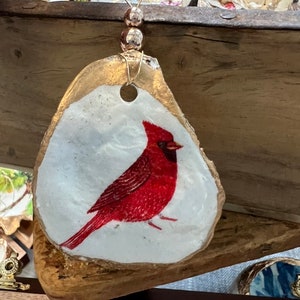 May include: A red cardinal painted on a white seashell ornament with gold accents. The ornament is attached to a gold wire with gold beads.