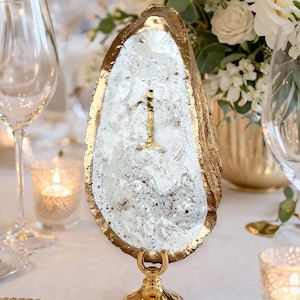 May include: A gold-edged oyster shell table number one, set on a gold stand. The shell has a white and speckled interior with a gold number one. The table setting includes white flowers, candles, and glassware.