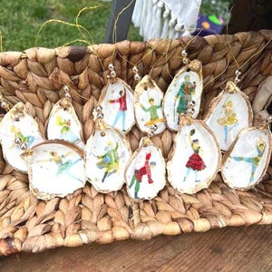 May include: A collection of 12 oyster shell ornaments, each featuring a different hand-painted illustration of a character from the Nutcracker ballet. The ornaments are strung with gold wire and beads, and are displayed in a woven basket.