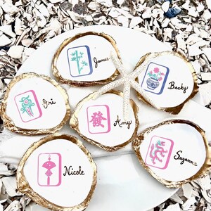 May include: White oyster shell place cards with gold edges, each featuring a different Mahjong tile design in pink and teal. Names are written in black script. A starfish rests on the plate. The shells are on a white plate, surrounded by white shells.