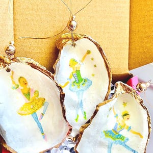 Op de afbeelding: Drie met goud geverfde oester schelp ornamenten met een witte achtergrond en geschilderde ballerina figuren. Elk ornament heeft een gouden touwtje om op te hangen.