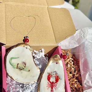 May include: Two oyster shell ornaments with gold trim. One ornament features a green alligator with red eyes and the other features a red lobster with green holly and red berries.