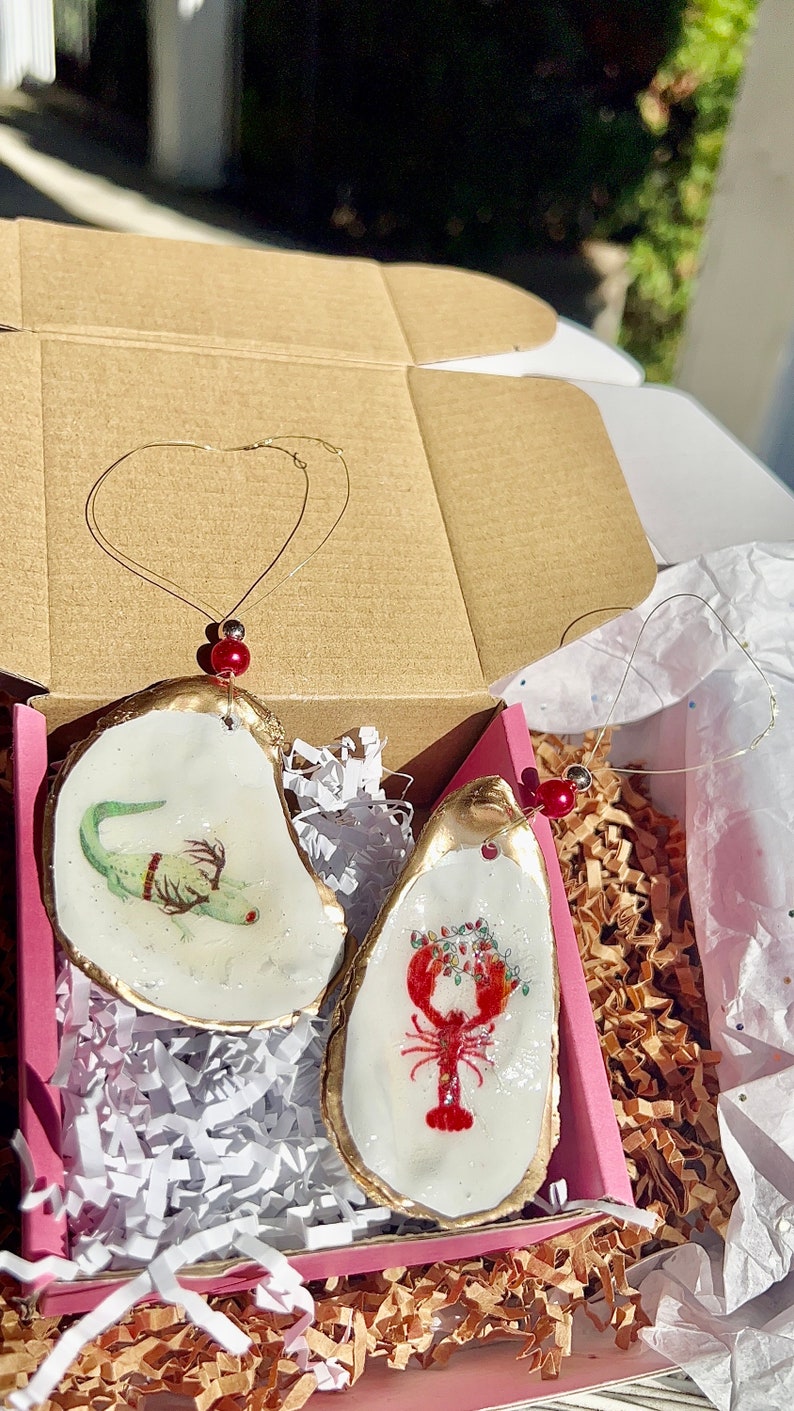 May include: Two oyster shell ornaments with gold trim. One ornament features a green alligator with red eyes and the other features a red lobster with green holly and red berries.