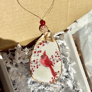 May include: A red cardinal bird ornament with gold accents and red berries on a white background. The ornament is in a white box with white shredded paper.