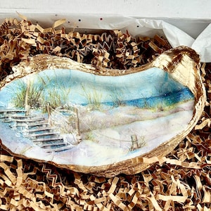 May include: A hand-painted oyster shell with a seascape scene. The scene depicts a wooden staircase leading down to a sandy beach with blue water and green grass in the background.