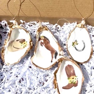 May include: Four oyster shell ornaments with gold trim and various designs. One features a brown horse, another a horseshoe, another a cowboy boot, and the last a hat with sunflowers.