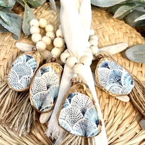 May include: Set of four blue and white oyster shell napkin rings with gold accents and jute tassels. The rings are strung on a white beaded garland.