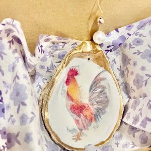 May include: A gold oyster shell ornament with a rooster illustration inside. The ornament is decorated with a white and purple floral pattern and has a small pearl hanging from it.