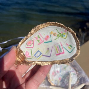 May include: A decorative oyster shell with a gold-painted rim, holding several small, colorful tiles with floral and geometric designs. The shell is held up against a blurred background of water. A starfish is visible in the lower right corner.