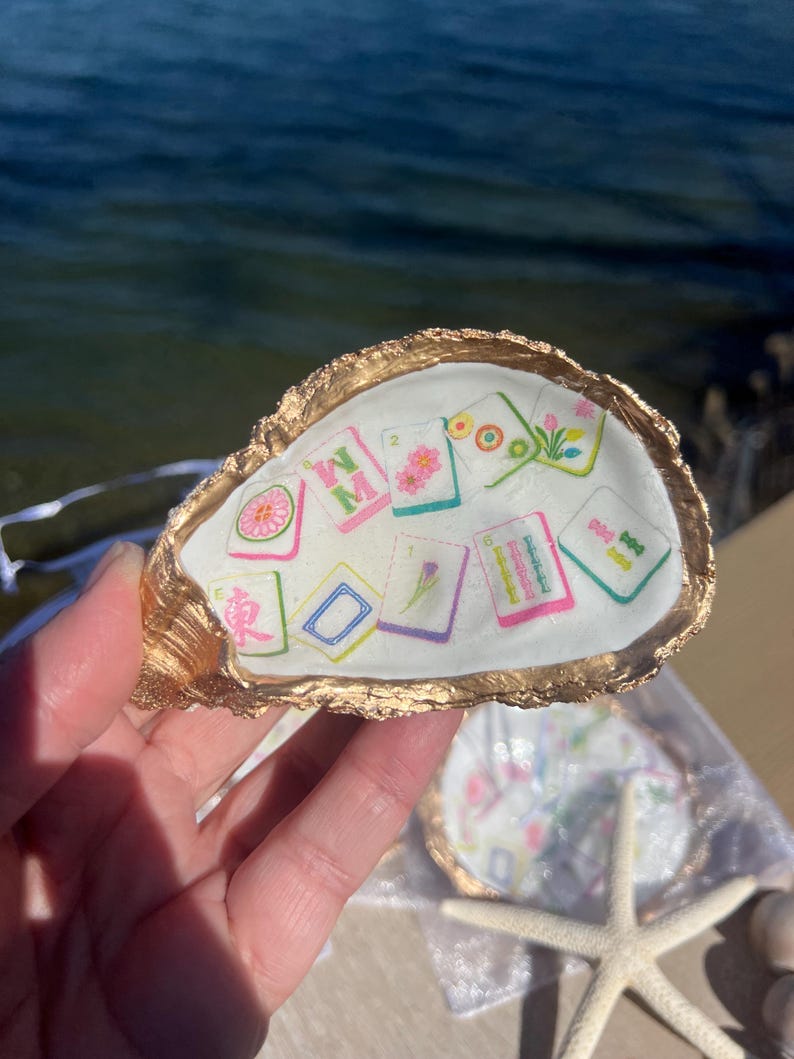 May include: A decorative oyster shell with a gold-painted rim, holding several small, colorful tiles with floral and geometric designs. The shell is held up against a blurred background of water. A starfish is visible in the lower right corner.