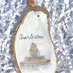 Op de afbeelding: Een witte oester schelp ornament met gouden rand, met een handgeschilderde illustratie van de Pineapple Fountain in Charleston, South Carolina. Het ornament is gelabeld "Charleston" in zwarte schrift.