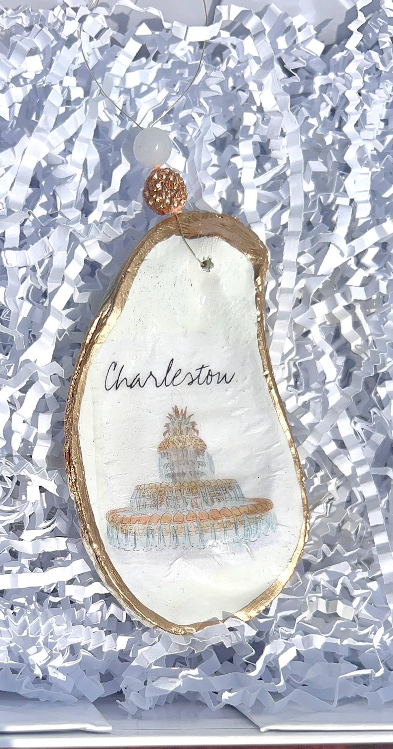 Op de afbeelding: Een witte oester schelp ornament met gouden rand, met een handgeschilderde illustratie van de Pineapple Fountain in Charleston, South Carolina. Het ornament is gelabeld "Charleston" in zwarte schrift.