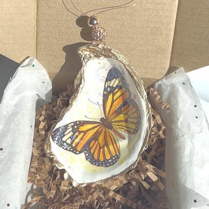 May include: A gold-painted oyster shell ornament with a hand-painted butterfly design. The butterfly has orange, yellow, and blue wings. The ornament is hanging from a gold wire with a small gold bead.
