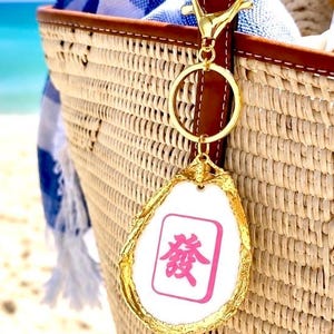 May include: A gold-colored keychain with a white and pink Mahjong tile charm, attached to a woven straw tote bag with a brown leather handle. The charm has a pink Chinese character. A blue and white striped towel is inside the bag.