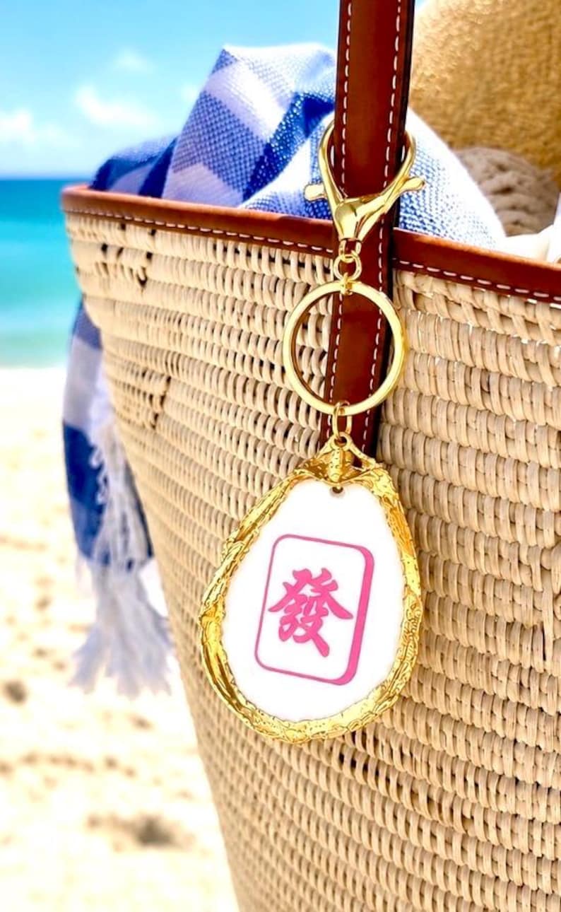 May include: A gold-colored keychain with a white and pink Mahjong tile charm, attached to a woven straw tote bag with a brown leather handle. The charm has a pink Chinese character. A blue and white striped towel is inside the bag.