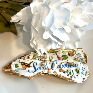 Op de afbeelding: Een witte keramische ringenschotel in de vorm van de staat South Carolina. De schotel is versierd met een kleurrijke kaart van de staat, inclusief bezienswaardigheden en de tekst "South Carolina". Twee gouden ringen liggen op de schotel.