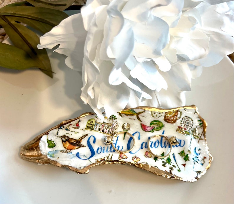 Op de afbeelding: Een witte keramische ringenschotel in de vorm van de staat South Carolina. De schotel is versierd met een kleurrijke kaart van de staat, inclusief bezienswaardigheden en de tekst "South Carolina". Twee gouden ringen liggen op de schotel.
