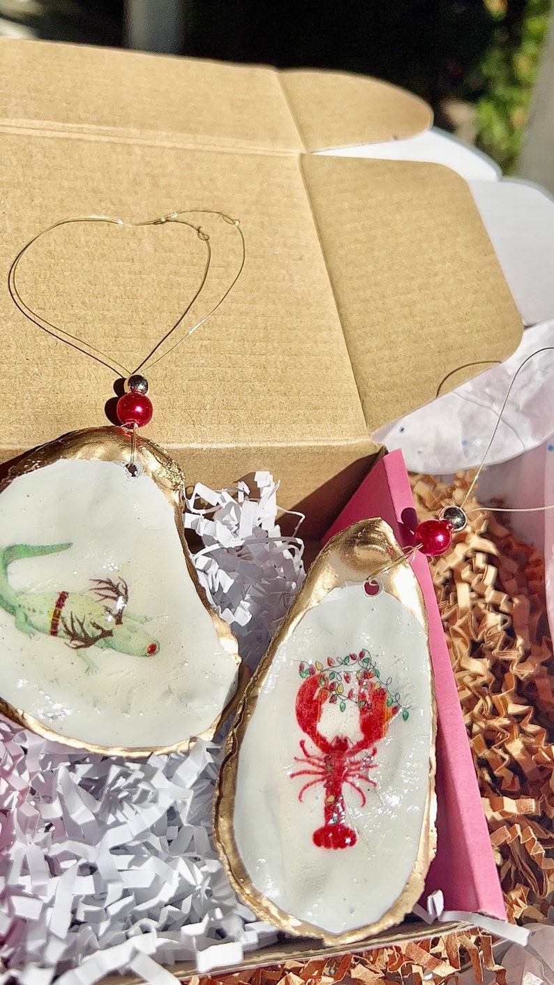 May include: Two oyster shell ornaments with gold trim. One ornament features a green lizard with a red and gold patterned band around its body. The other ornament features a red lobster with a garland of red and green lights around it.