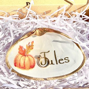 Puede incluir: Un plato decorativo de concha marina pintado a mano. El plato presenta un borde dorado y una calabaza naranja pintada con hojas de otoño. El nombre "Jules" está escrito en letras doradas. Un anillo dorado está colocado en el plato.