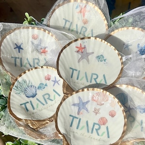 May include: White seashell-shaped coasters with gold trim, featuring starfish, seashells, and the text "TIARA on the beach" in teal. The coasters are packaged in clear bags, perfect for a beach-themed event.