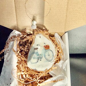 May include: A white oyster shell ornament with a painted image of a light blue bicycle with a basket of flowers. The ornament is packaged in a white box with brown paper packing material.