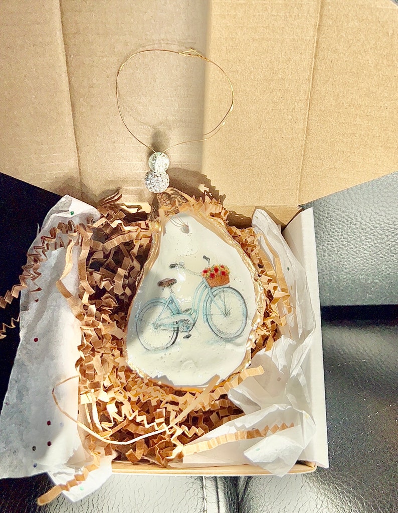 May include: A white oyster shell ornament with a painted image of a light blue bicycle with a basket of flowers. The ornament is packaged in a white box with brown paper packing material.