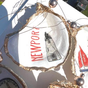 Pode incluir: Um conjunto de três conchas de ostra com borda dourada e designs diferentes. A concha superior tem uma ilustração de um farol e o texto "NEWPORT" em vermelho. A concha do meio tem uma âncora náutica e o texto "LEN'S WHARF" em preto. A concha inferior tem uma ilustração de um veleiro e o texto "NEWPORT" em vermelho.